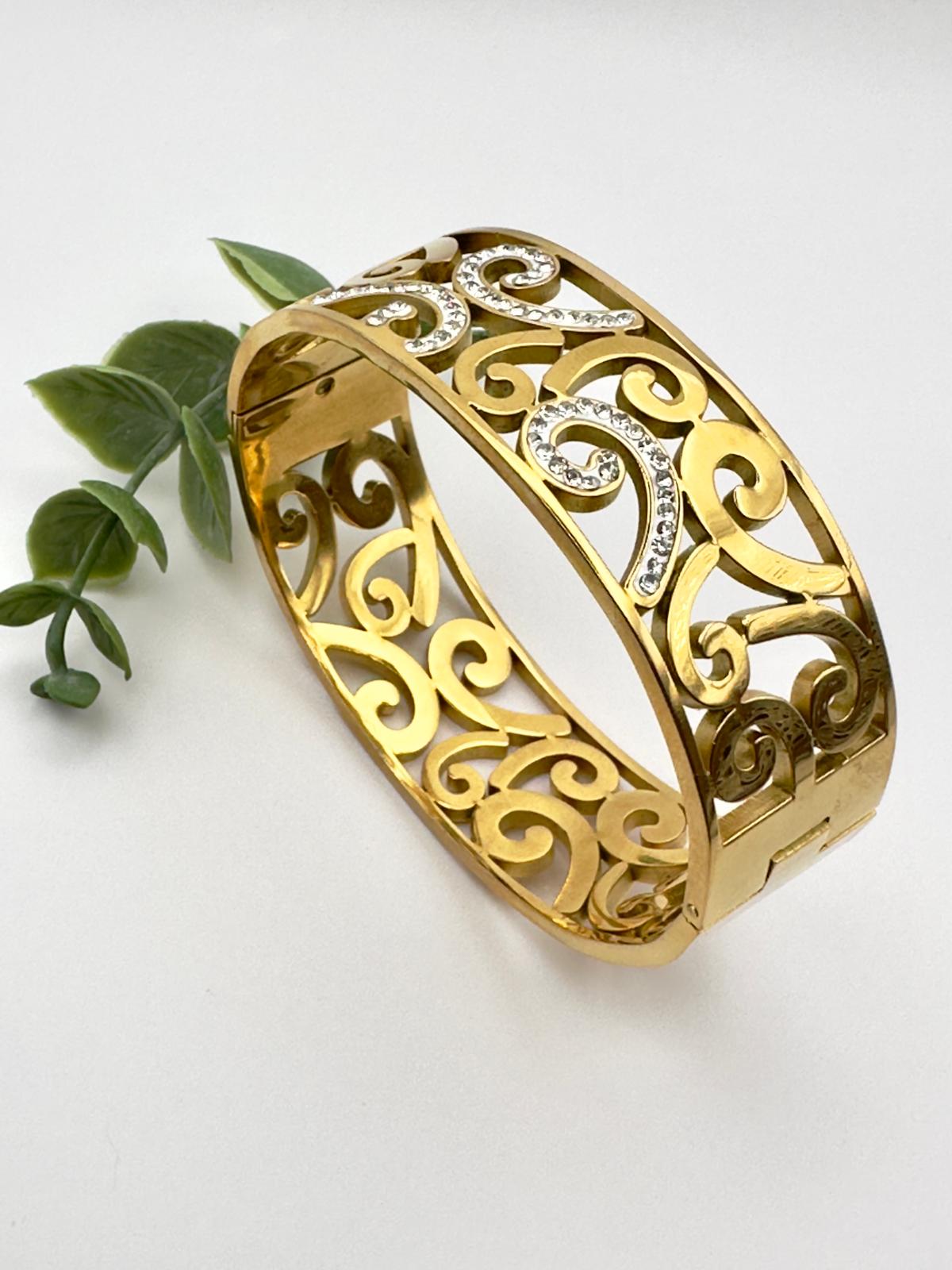 May include: Gold-toned hinged bangle bracelet with an openwork design of swirling patterns. The bracelet features clear, sparkling accents and a secure clasp. A stylish piece of jewelry.